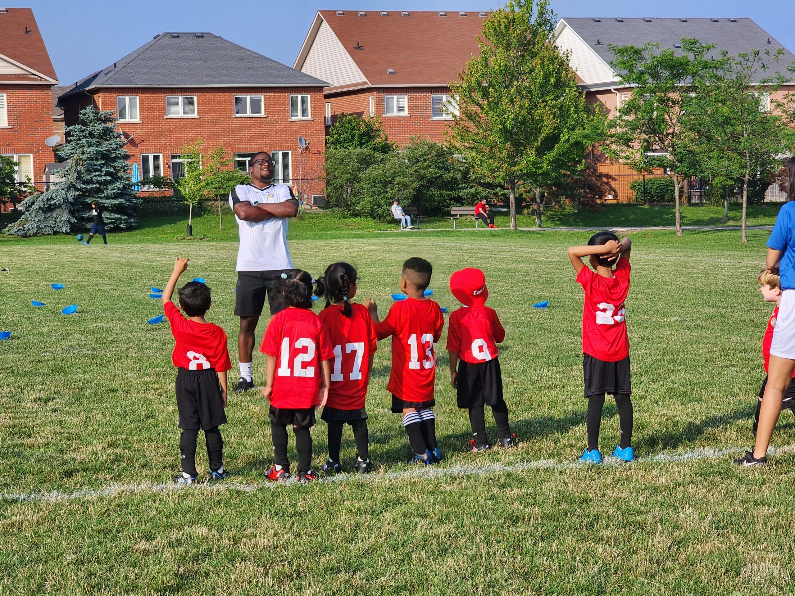 Young children aged 4–6 dribbling and passing a soccer ball on a grassy field under the guidance of a coach, all smiling and actively engaged in play.